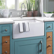 The Astini Belfast 100 white ceramic kitchen sink with drainer and strainer waste is set in blue cabinets with wood accents, featuring a striped dish towel, two potted plants, and a bowl of greens by the window above.